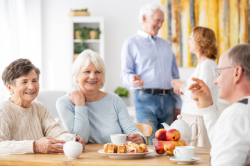Lächelnde Personen unterschiedlichen Alters sitzen oder stehen am Tisch mit Kuchen und Kaffee
